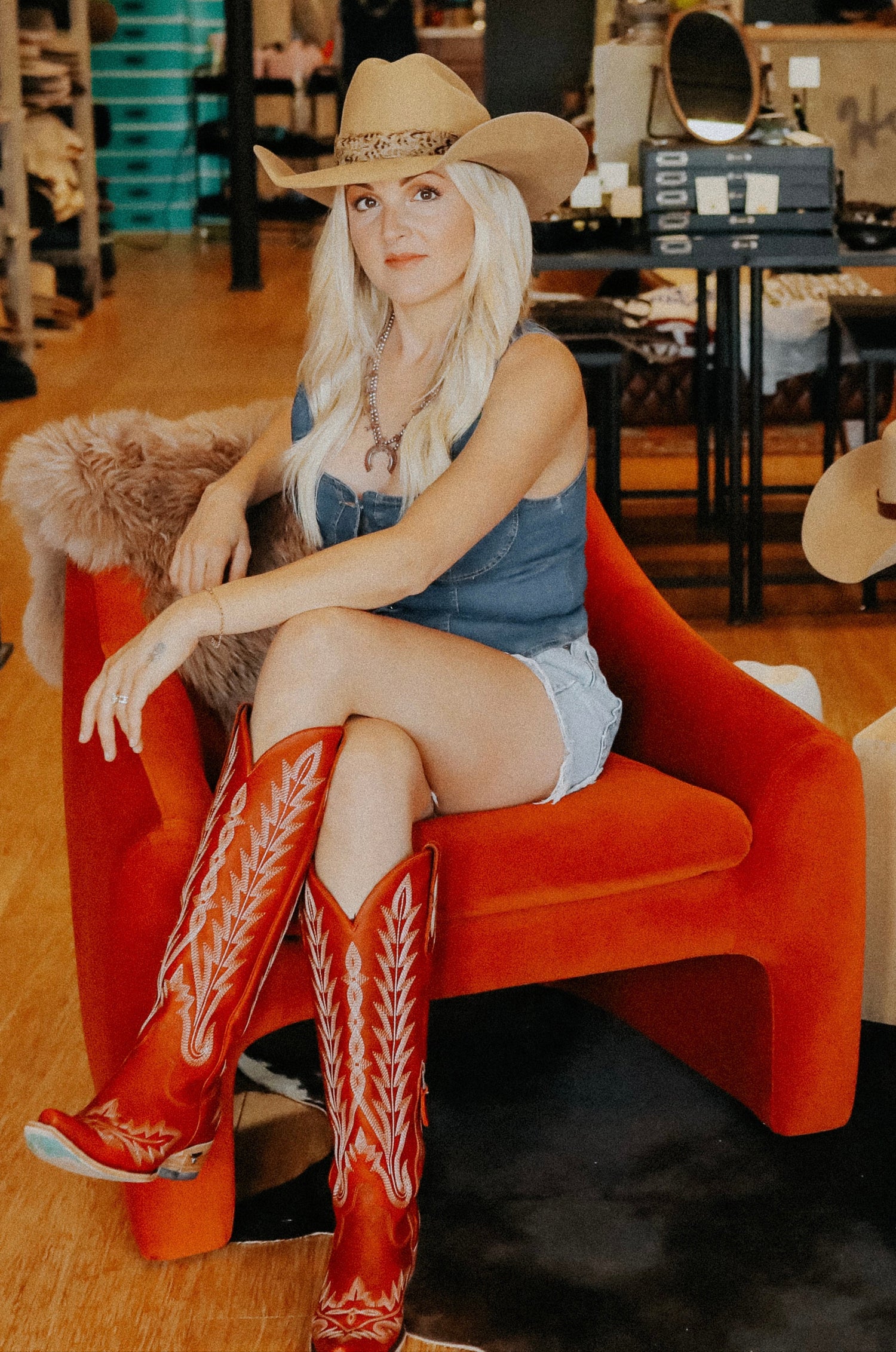 Woman in a store wearing a cowboy hat and boots, sitting on an orange chair.